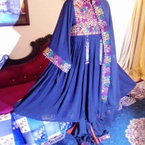 A Blue Velvet Afghani Frock with vibrant hand embroidery in floral and geometric patterns across the chest, sleeves, and collar, displayed elegantly on a red velvet sofa. The intricate detailing features shades of pink, green, yellow, and white, highlighting traditional Afghan craftsmanship. The room setting includes a blue-patterned carpet, framed wall art, and soft curtain lighting, creating a regal backdrop that enhances the frock’s cultural beauty