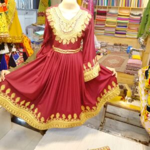 Maroon Afghani Frock with intricate gold embroidery, long sleeves, and flared skirt, displayed on a mannequin in a traditional fabric shop with colorful textiles in the background.