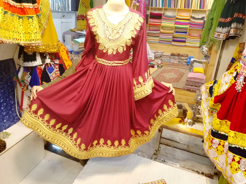 Maroon Afghani Frock with intricate gold embroidery, long sleeves, and flared skirt, displayed on a mannequin in a traditional fabric shop with colorful textiles in the background.