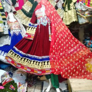 Maroon velvet Afghani frock with multicolor embroidery and golden threadwork, displayed on a mannequin with a sheer red dupatta and surrounded by traditional Afghan garments in a cultural boutique.