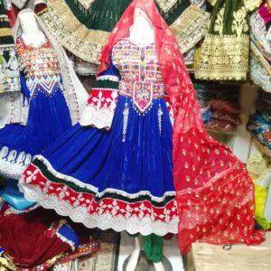 "Blue Velvet Afghani Frock adorned with vibrant traditional embroidery in red, white, and green, featuring a full skirt with white lace hem and intricate border detailing. A red dupatta with golden accents complements the rich design. Set against a backdrop of cultural garments, the frock showcases Afghan heritage and handcrafted artistry."