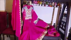 "A vibrant pink Afghan dress with colorful floral embroidery on the bodice and sleeves, displayed on a chair with a matching pink dupatta. The outfit is arranged to highlight its traditional craftsmanship, with a background shelf holding small decorative pots and plants."