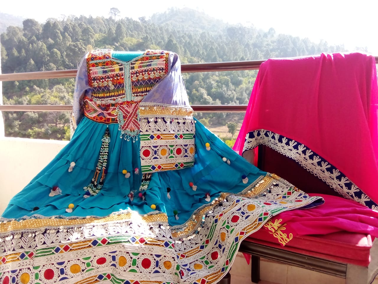 A traditional Afghan dress in deep blue with intricate multicolored embroidery, paired with a vibrant pink shawl featuring decorative borders. The outfit is displayed on a chair placed on a balcony, with a scenic view of green trees and distant mountains in the background. This image reflects Afghan embroidery storytelling, where each stitch conveys cultural heritage and artisanal elegance.