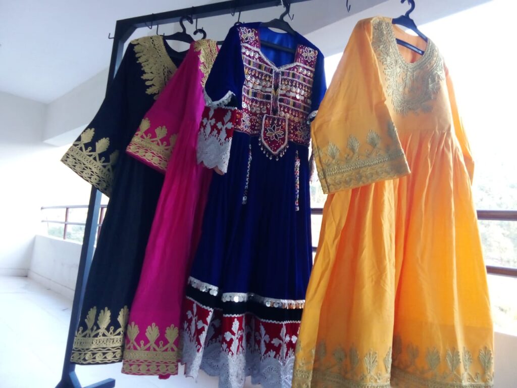 A selection of Velvet Embroidered Frocks displayed on a rack, featuring four traditional dresses in black, pink, blue, and yellow. Each frock showcases intricate gold and white embroidery, reflecting cultural elegance and festive style.
