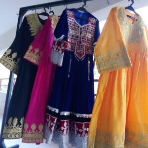 A selection of Velvet Embroidered Frocks displayed on a rack, featuring four traditional dresses in black, pink, blue, and yellow. Each frock showcases intricate gold and white embroidery, reflecting cultural elegance and festive style.