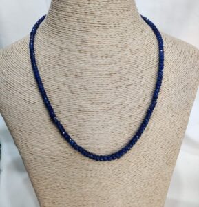 A necklace made of deep blue faceted beads elegantly displayed on a beige woven mannequin bust. The beads reflect light softly, creating a subtle shimmer against the neutral background. The design is simple yet refined, showcasing the rich sapphire color prominently.