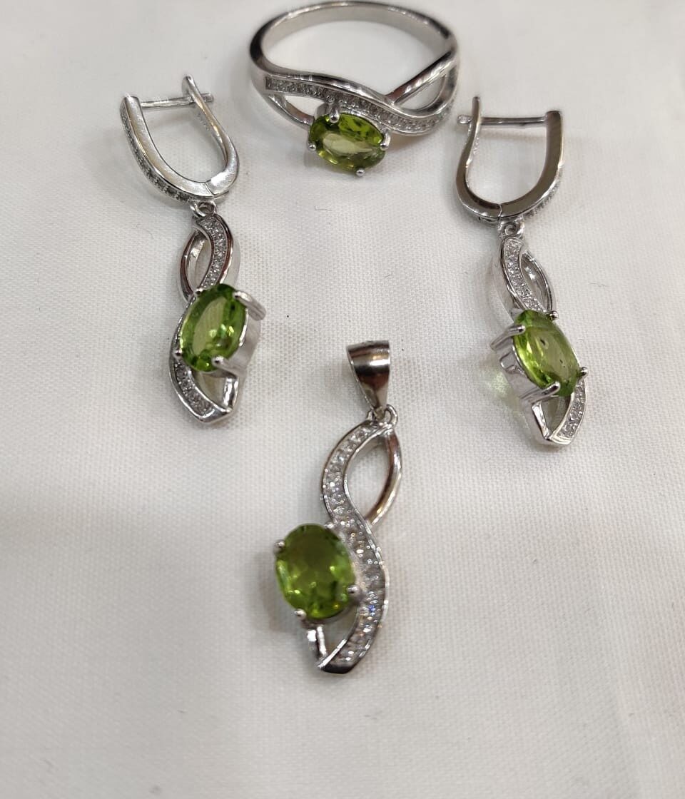 Silver peridot jewelry set displayed on soft white fabric, including a ring, pendant, and earrings with green gemstones. 