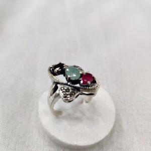 Silver ring with green and red gemstones, elegantly set in an intricate design, displayed atop a white circular stand against a soft gray background. The account page